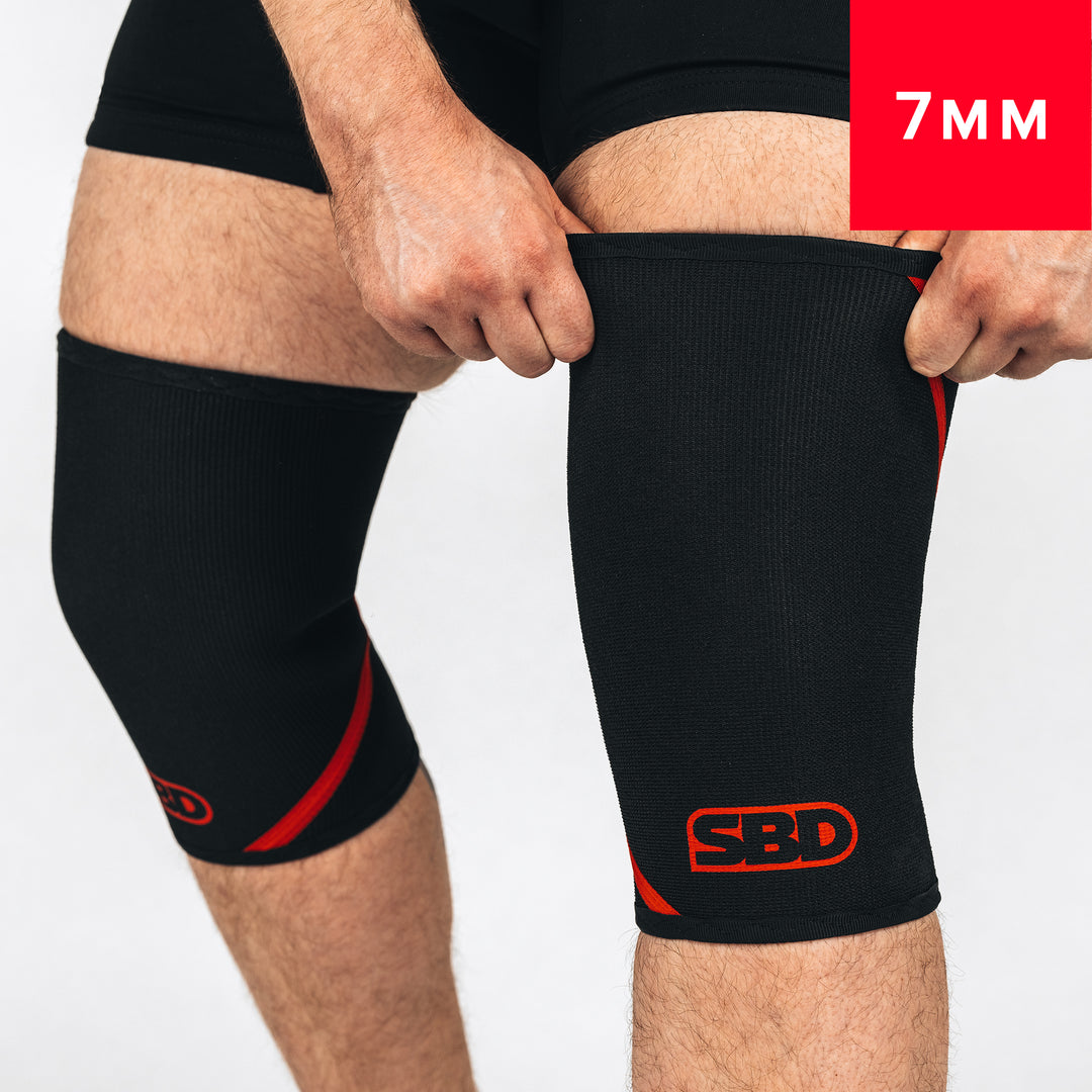 SBD Powerlifting Knee Sleeves 7mm – zwart-rode sleeves worden opgetrokken, met 7mm-aanduiding