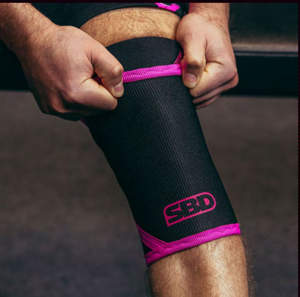 SBD Powerlifting Knee Sleeves 7mm - Nova Limited Edition – zwarte sleeve met felroze bies wordt rond de knie aangetrokken