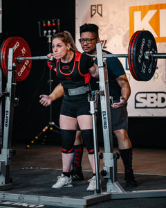 SBD Powerlifting Singlet Classic – rood-zwarte uitvoering gedragen door een vrouw tijdens een squat
