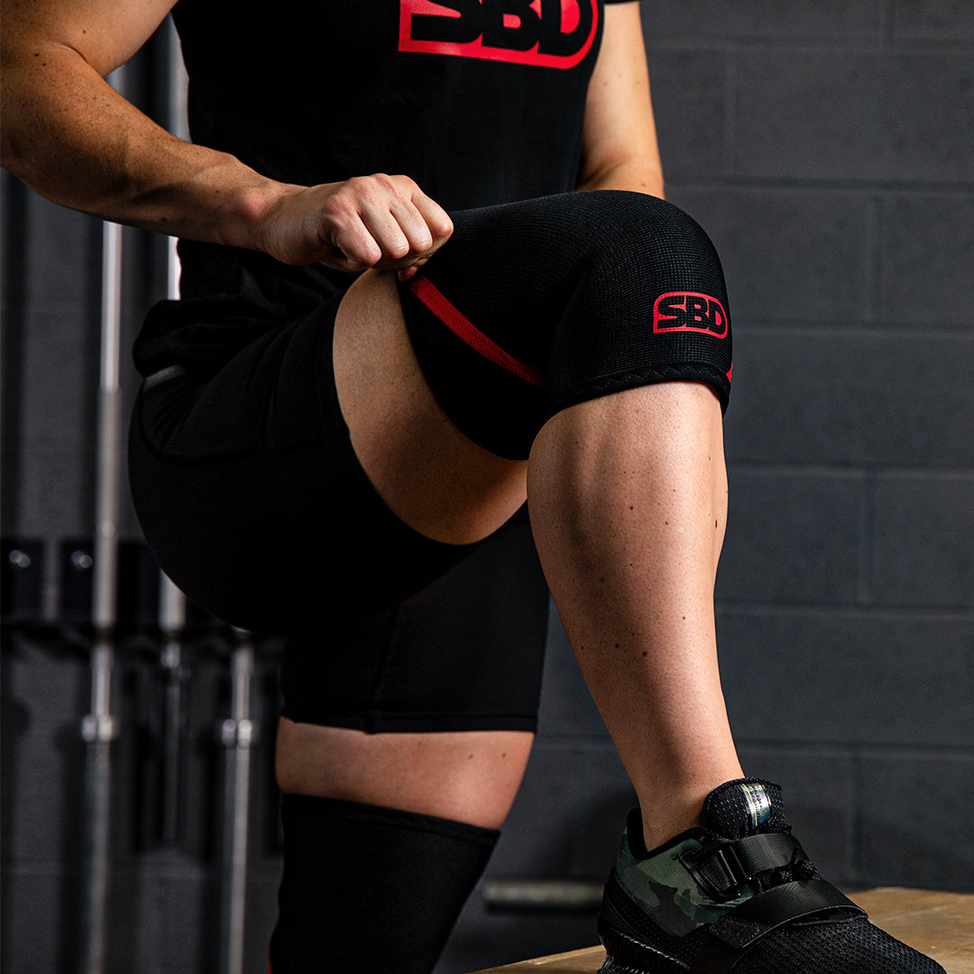 SBD Weightlifting Knee Sleeves 5mm – zwart-rode sleeves gedragen terwijl de atleet op een bank zit